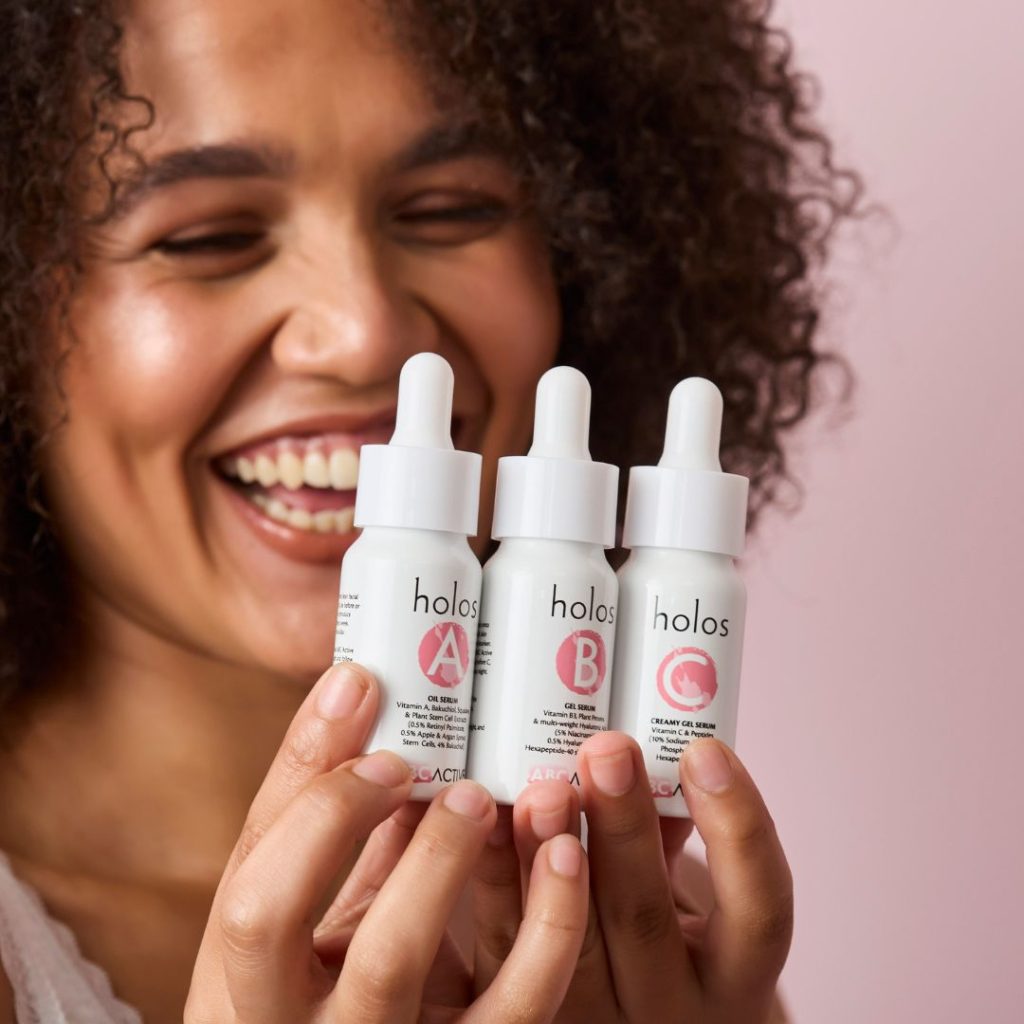 A smiling woman with curly hair holds three white dropper bottles labeled "holos" with large letters A, B, and C in pink circles. These bottles contain the Holos ABC Active range of vitamin serums, and the background is a soft pastel pink.