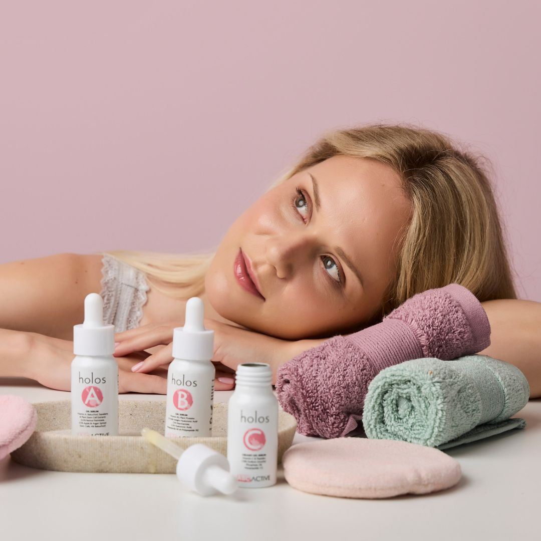 A woman with blonde hair rests her head on her arms on a table with a pink backdrop. In front of her are three skincare serum bottles labeled A, B, and C from the Holos ABC Active range vitamin serums, a stone dish with a dropper, a pink facial cloth, and rolled towels.